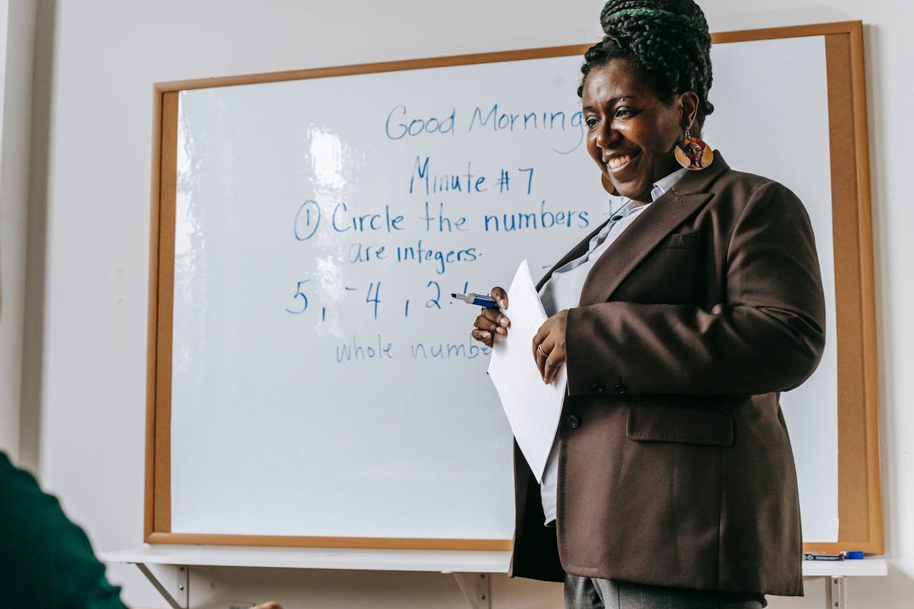 services-01 Crop smiling African American female teacher standing near whiteboard with papers in hands during math lesson
